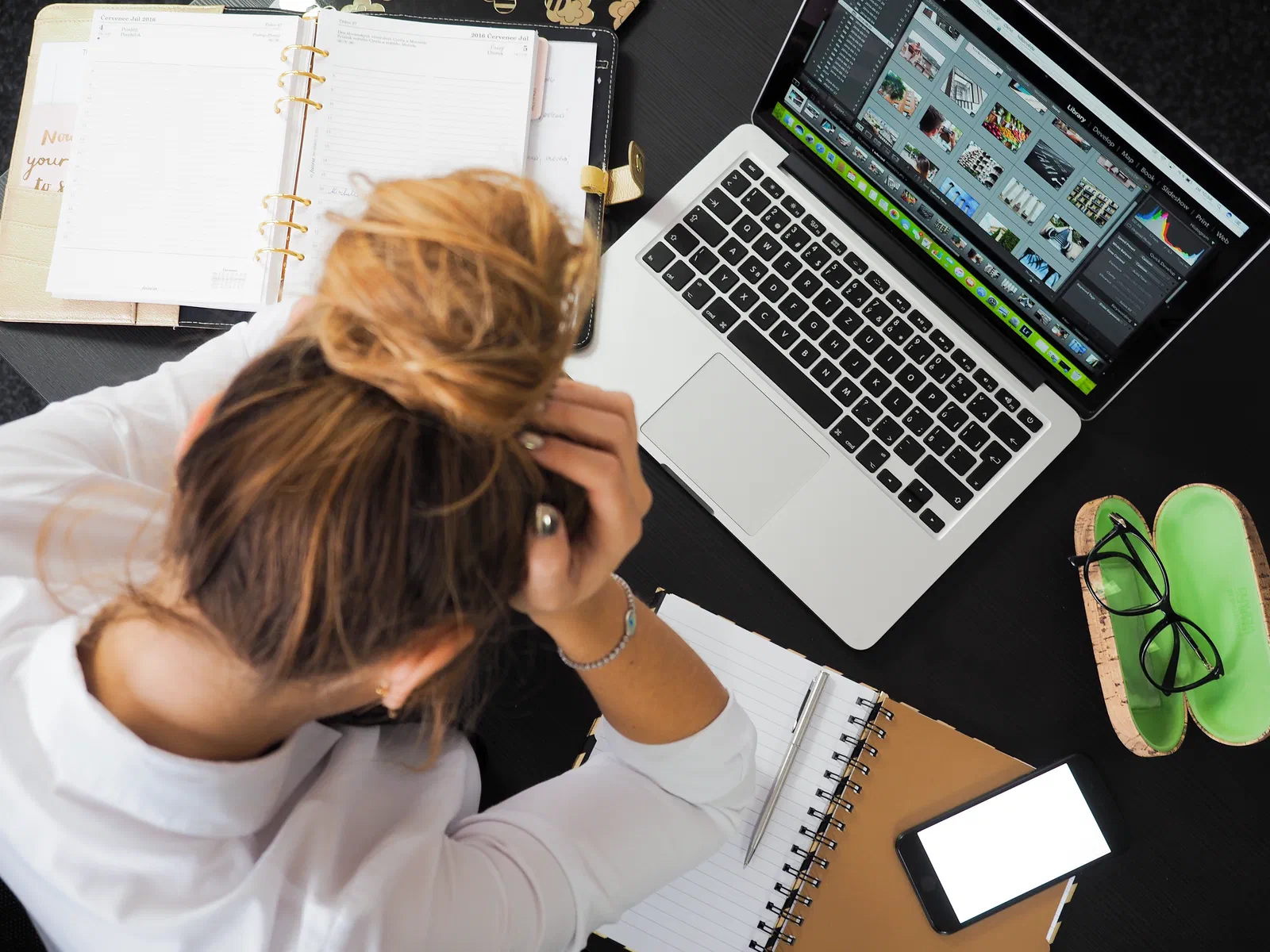 A stressed woman at her desk holding her head in her hands in front of a laptop and notebooks